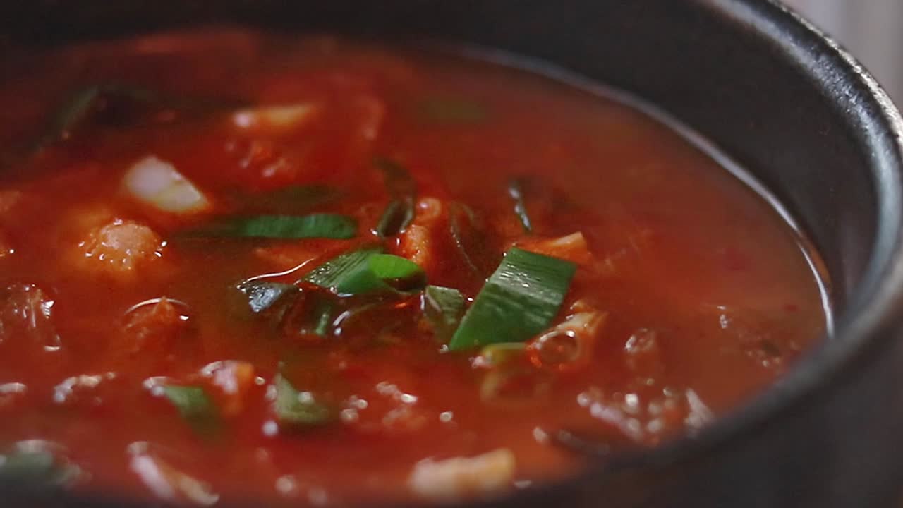 Close-up of a bubbling simmering red soup with green onions in a black bowl called kimchi jjgae or kimchi stew, authentic traditional local South Korean food