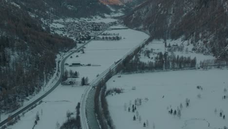 Red-Swiss-train-riding-through-snowy-valley-next-to-river-in-shadow-on-sunny-winter-morning-with-lush-pine-forests-on-hillsides-in-Swiss-Alps-with-small-picturesque-village-in-background