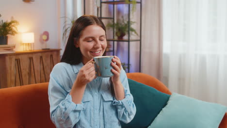 attractive smiling young woman girl drinking a cup of coffee or herbal tea sitting on home couch