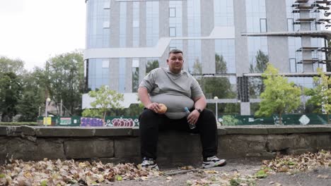 a fat man is holding a hamburger and water while taking a rest during his morning workout in a city a fat man is holding a hamburger and water while taking a rest during his morning workout in a city park the problem of excess weight in a young guy motivation to do sports