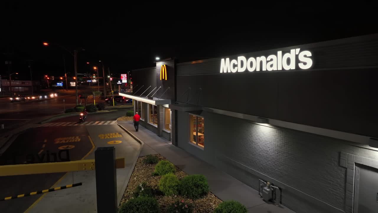 Person walking on sidewalk of Mc Donalds Restaurant at night. Famous fast food building in USA. Motorbike entering drive thru line. Aerial establishing shot.