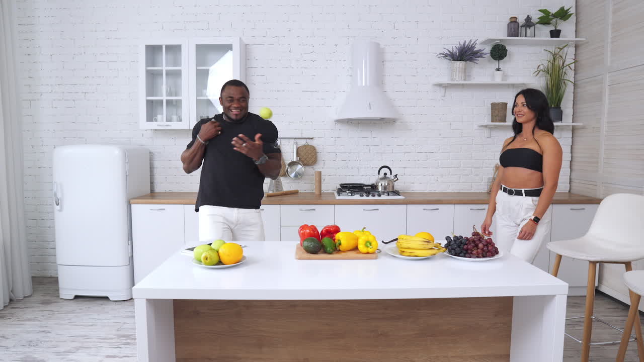 Diverse couple having good time in the home kitchen. Muscular Afro-American man trying to juggle with green apples. Caucasian girl watching him and smiling.