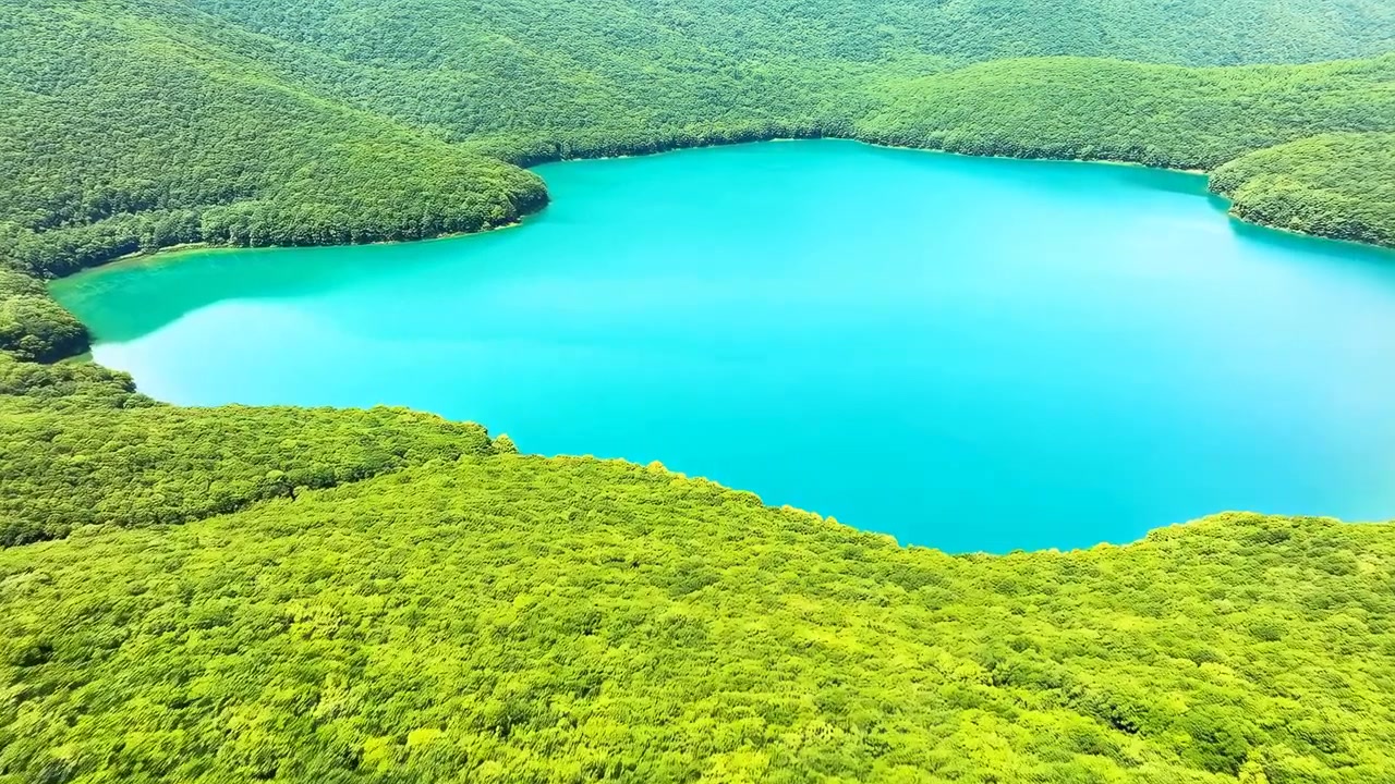 Høy skrå flyfoto som viser en turkis innlandssjø omgitt av tett eviggrønn skog, bølgende åser og ulendt kystlinje, med glatt blått vann, solfylte sommertoner og rik vegetasjon.