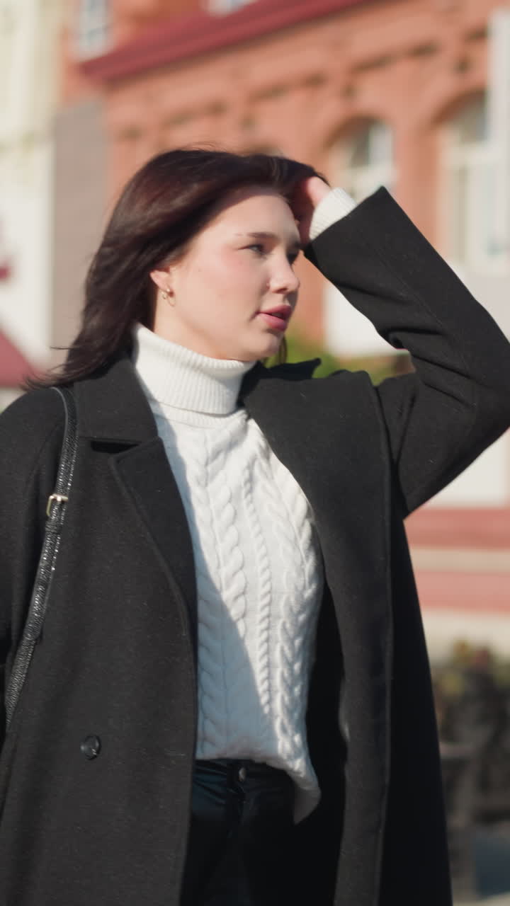 검은 코트와  스웨터를 입은 러시아 여성은 검은 드백을 들고 왼쪽으로 보면서 머리를 조정합니다. 배경에는 흐릿한 주거 건물이 있습니다.