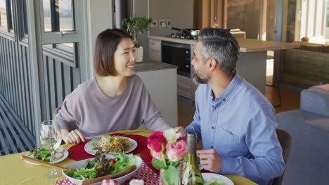 happy diverse couple sitting at table in dining room, eating dinner and drinking wine