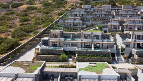 Vista-De-Cayoresort-En-Grecia-Creta---Mujer-De-Pelo-Moreno-En-Traje-De-Baño-Sentada-Junto-A-La-Piscina-Y-Bebiendo-Un-Cóctel---Drone-Volando-Hacia-Atrás-Rápidamente