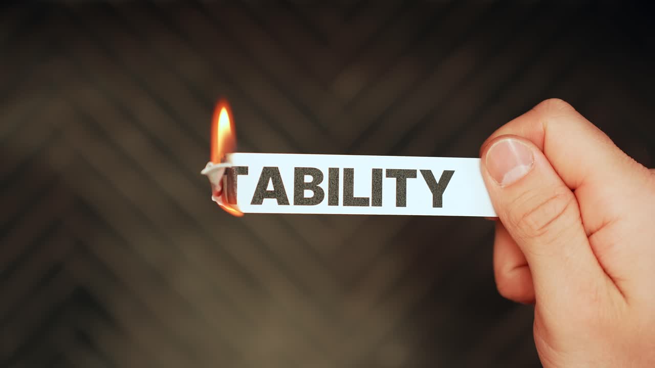 Close-up of a hand holding a paper strip labeled “STABILITY” as the edge burns with flame on a dark background. Economic uncertainty and life instability concept with copy space
