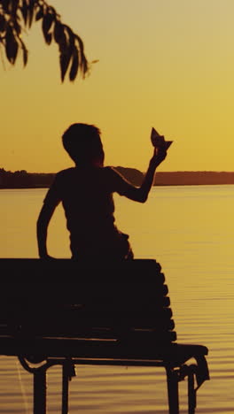 a little boy is sitting on the bench and playing with a paper boat on the background of evening landscape by the lake in the summer. sunset vertical video