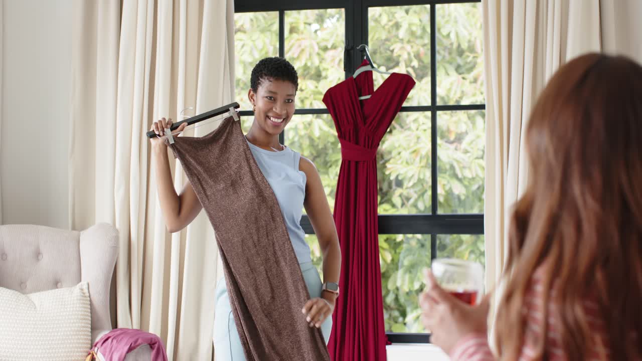 Amigas de diversas etnias observando a una mujer afroamericana que sostiene un vestido marrón en el dormitorio, pidiendo opinión.