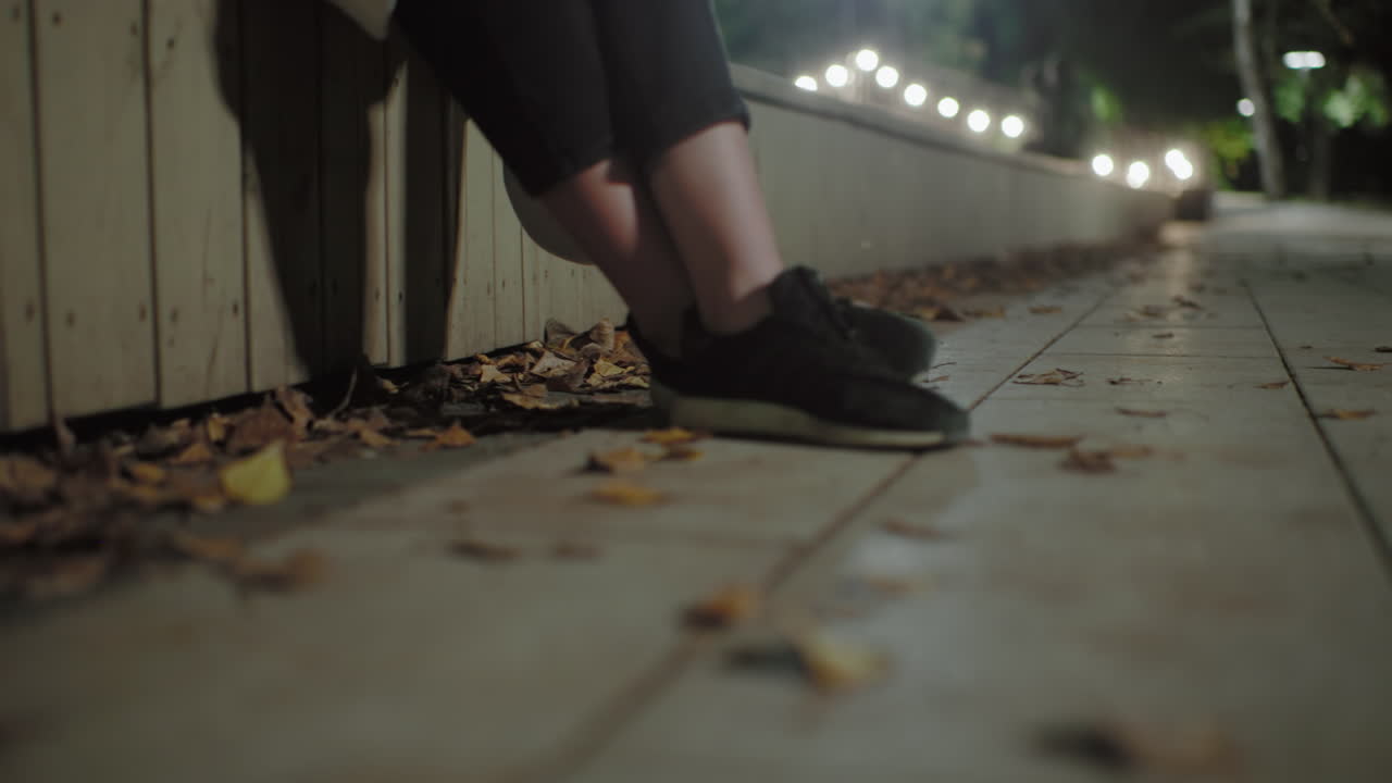 Close up of crossed legs of seated individual in black sneakers resting on dried autumn leaves along tiled pathway, blurred bokeh street lamps and trees create calm nighttime park atmosphere