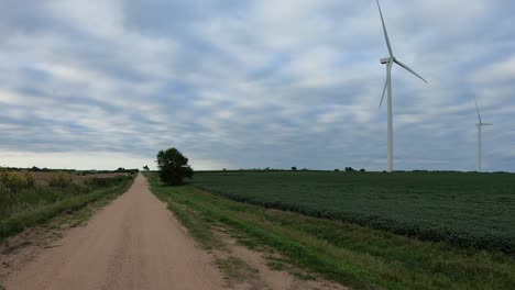 Auf-Einer-Ländlichen-Schotterstraße,-Die-An-Einem-Bewölkten-Tag-Eine-Drehende-Windkraftanlage-Beobachtet,-Die-Sich-In-Einem-Sojabohnenfeld-Entlang-Einer-Schotterstraße-Im-Ländlichen-Nebraska-Usa-Befindet