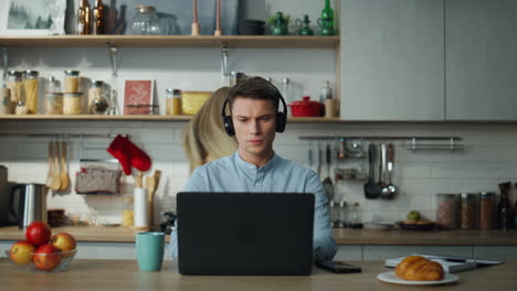 hombre escuchando auriculares de cursos en línea sentado en la mesa de la cocina bebiendo café.