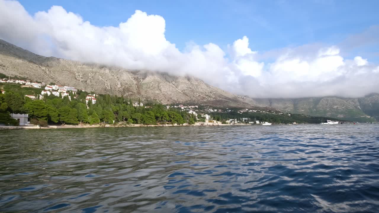 A static wide shot of a Croatia Coastline scene. Ocean in the foreground with trees on the farside shore. Then mountains behind and some clouds in the sky