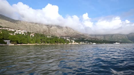 a static wide shot of a croatia coastline scene. ocean in the foreground with trees on the farside shore. then mountains behind and some clouds in the sky