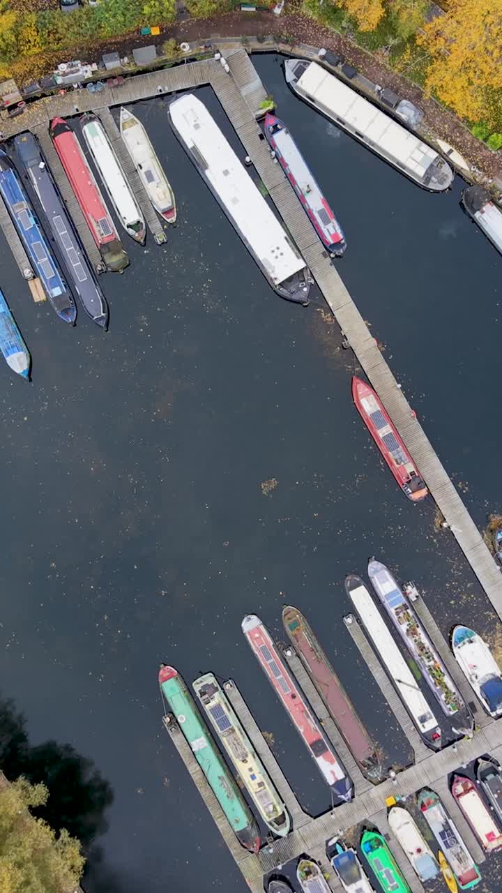 Vertical high drone orbit over Harlow Marina, Essex. The top-down view shows colourful narrowboats and autumn trees. The still water reflects leaves and debris, creating a calm seasonal aerial scene