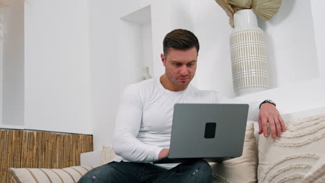 concentrated caucasian male sitting on sofa looking at laptop. man in white shirt works at home remote from office.