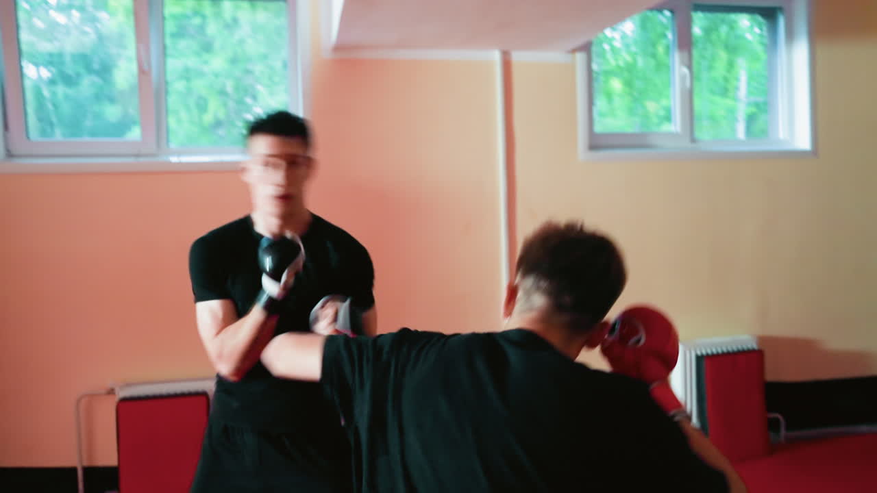 Fighters engaged in sparring session inside gym, one delivering punch while other bends forward, both wearing gloves and black shirts, demonstrating action, coordination, athletic combat training
