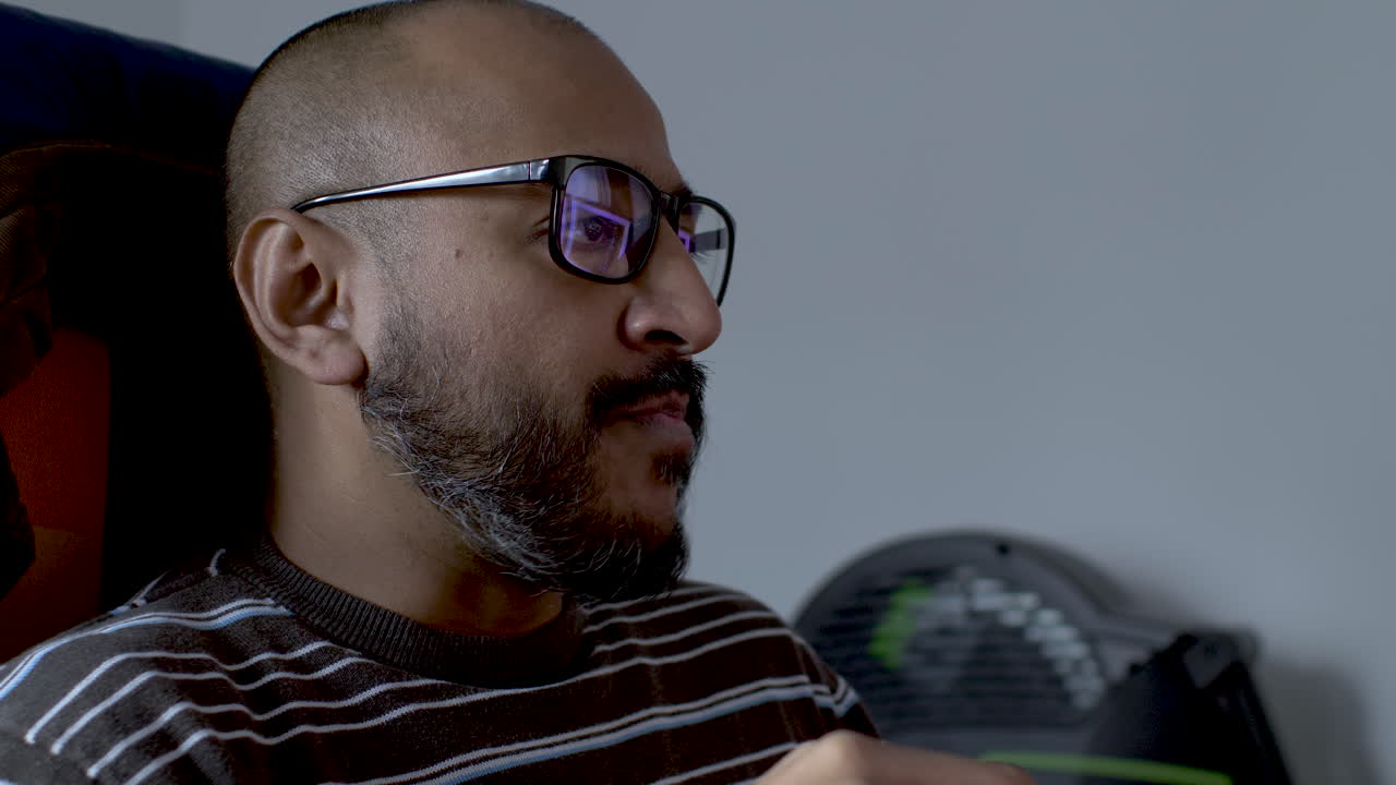 A close up shot of the face of a casually dressed Indian man focused on his computer screen reading while taking the opportunity to munch on a quick food snack