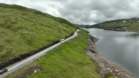 Aerial-following-white-bus-and-black-car-along-small-lake-at-Vikafjell-mountain-pass-in-Norway-during-summer