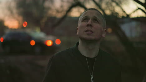 a man in a green jacket stands outdoors at dusk, smoking a cigarette, with bokeh lights from distant cars and surrounding trees