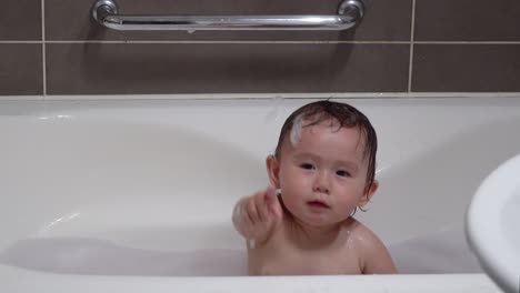 mixed asian toddler baby girl clapping hands while bathing in a foamy bathtub in the bathroom