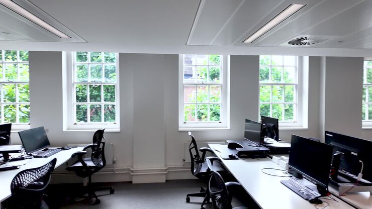 A bright, contemporary empty office space featuring multiple workstations, ergonomic chairs, and large windows allowing natural light to illuminate the room