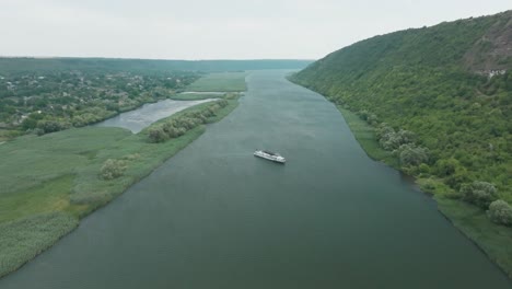 boat-that-is-slowly-moving-in-the-middle-of-the-river,-located-to-a-rural-type-settlement-on-the-other-side---this-is-the-edge-of-the-mountain-green-trees-grow-on-the-edge-and-a-lot-of-reeds