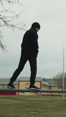 a young boy dressed in a black outfit carefully walks along an iron pole carefully, while another person, dressed in a gray jacket and beanie, rests against the pole, looking up