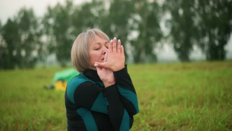 woman in green and black suit seated in cow face pose practicing yoga outdoors in a grassy field, hands gracefully raised, with yellow boots and green raincoat visible in the blurred background