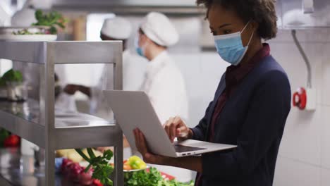 Portrait-of-african-american-female-manager-wearing-face-mask-using-laptop-in-restaurant-kitchen