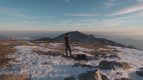 vista posterior de gran angular de un joven excursionista tomando fotos con su cámara en la cima de una montaña nevada en las montañas de guadarrama, madrid, españa