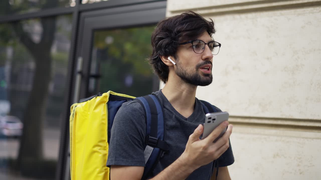Delivery man with yellow backpack talk to a client on the street using earphones