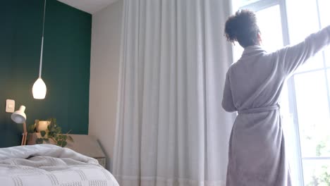 Back-of-biracial-woman-in-bathrobe-opening-curtains-in-bedroom,-slow-motion,-copy-space