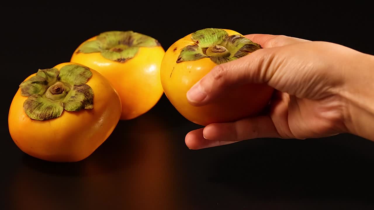 Close-up of hands handling and arranging ripe persimmons on a dark background.