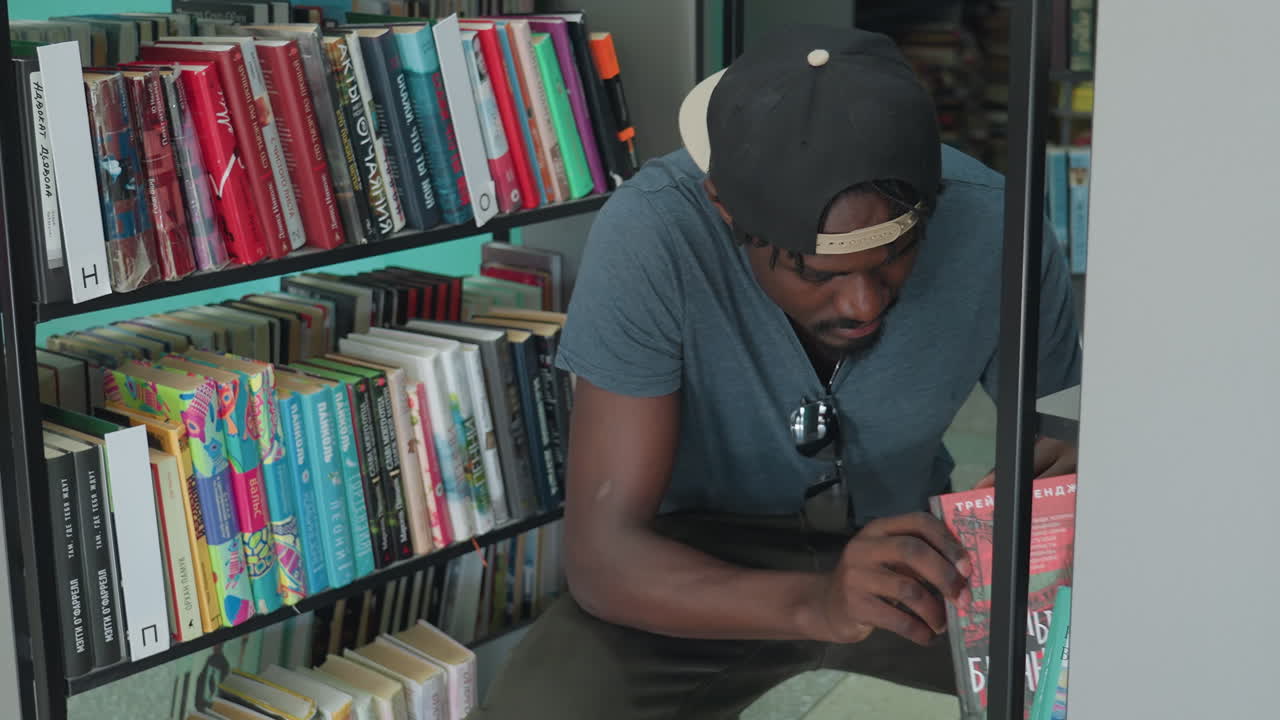 Young man in grey shirt and backwards cap squatting near library shelf, reaching to retrieve book while surrounded by colorful spines, focused on selecting reading material in quiet indoor setting