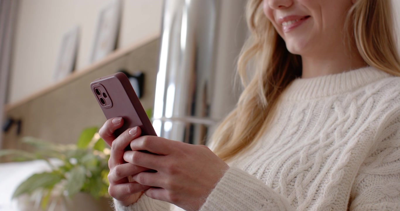 Donna con maglione color crema tiene in mano uno smartphone con custodia viola e legge i messaggi con i pollici