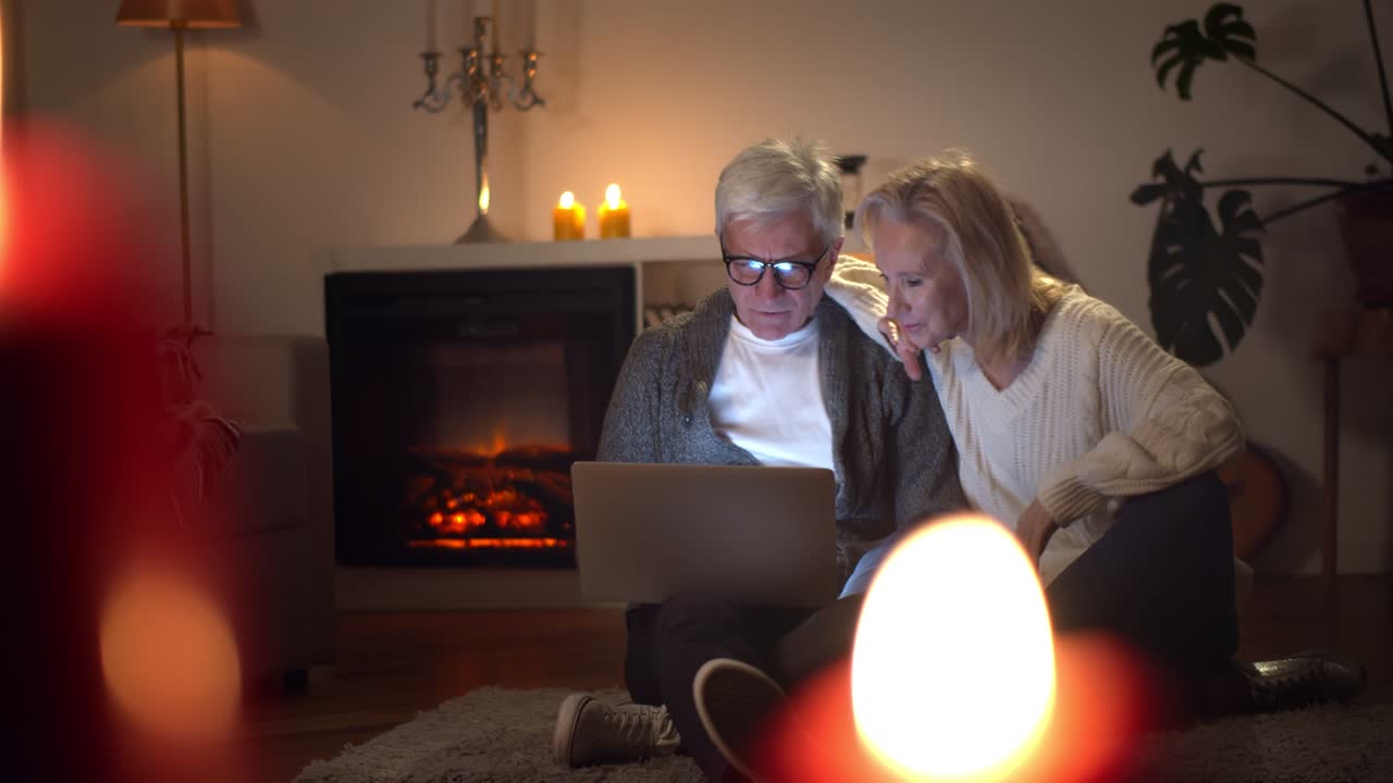 una pareja de ancianos navegando por internet juntos sentados cerca de la chimenea en el suelo