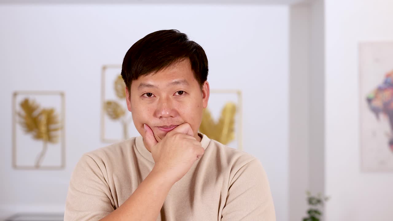 Asian man stands indoors, thoughtfully touching his chin while making various contemplative facial expressions. Soft, even lighting and steady camera create a calm mood