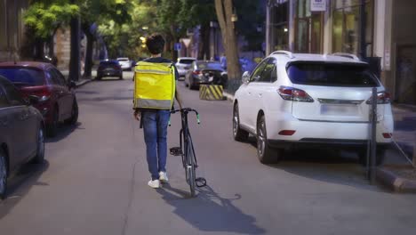 Delivery-man-walking-alone-with-bike-and-yellow-bag-by-evening-city,-rear-view