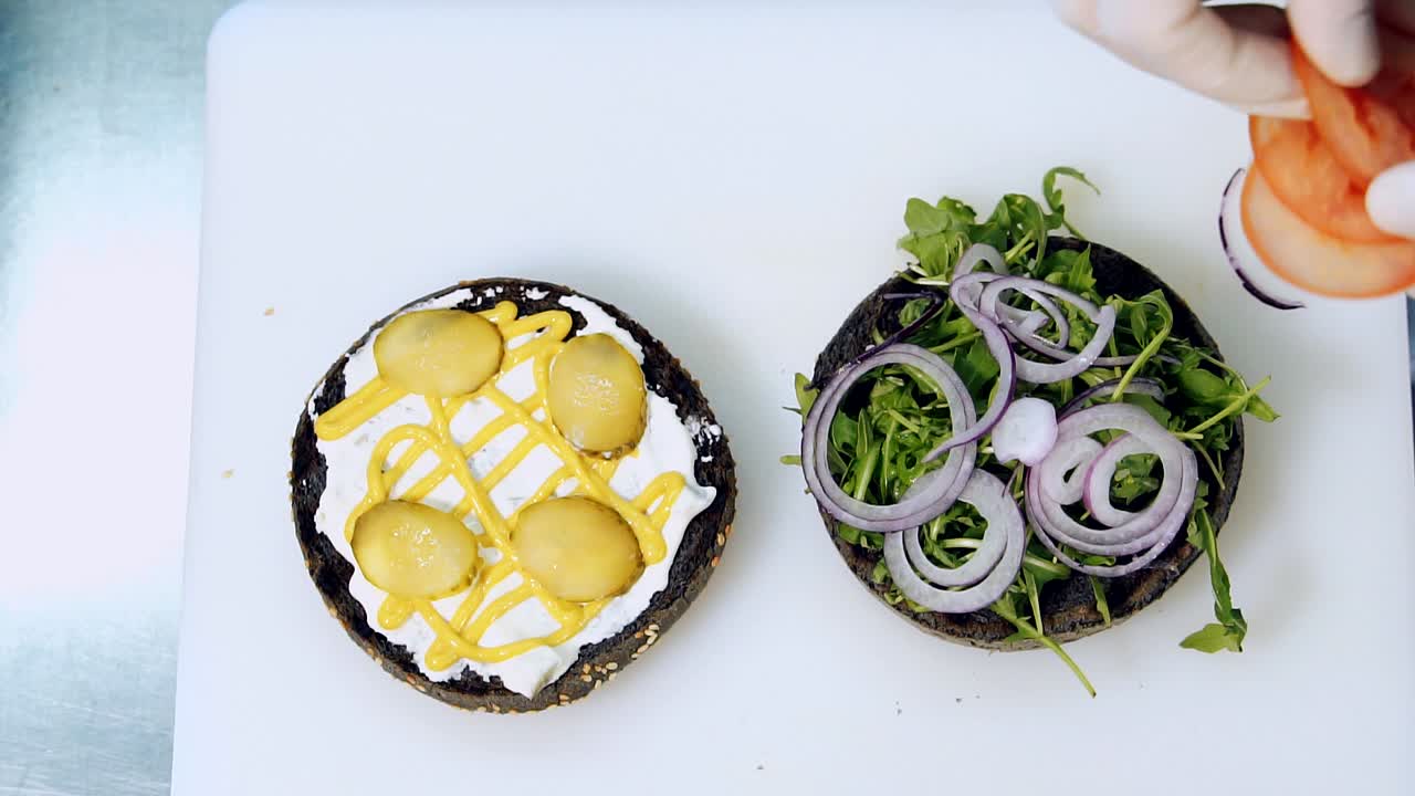 Cooking vegan burger. Chef putting fresh vegetables on black bun for making healthy hamburger. Cook puts tomato in burger. Delicious food for vegetarians