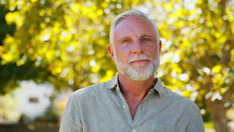 Portrait-Of-Smiling-Senior-Man-Standing-Outdoors-In-Garden-Park-Or-Countryside