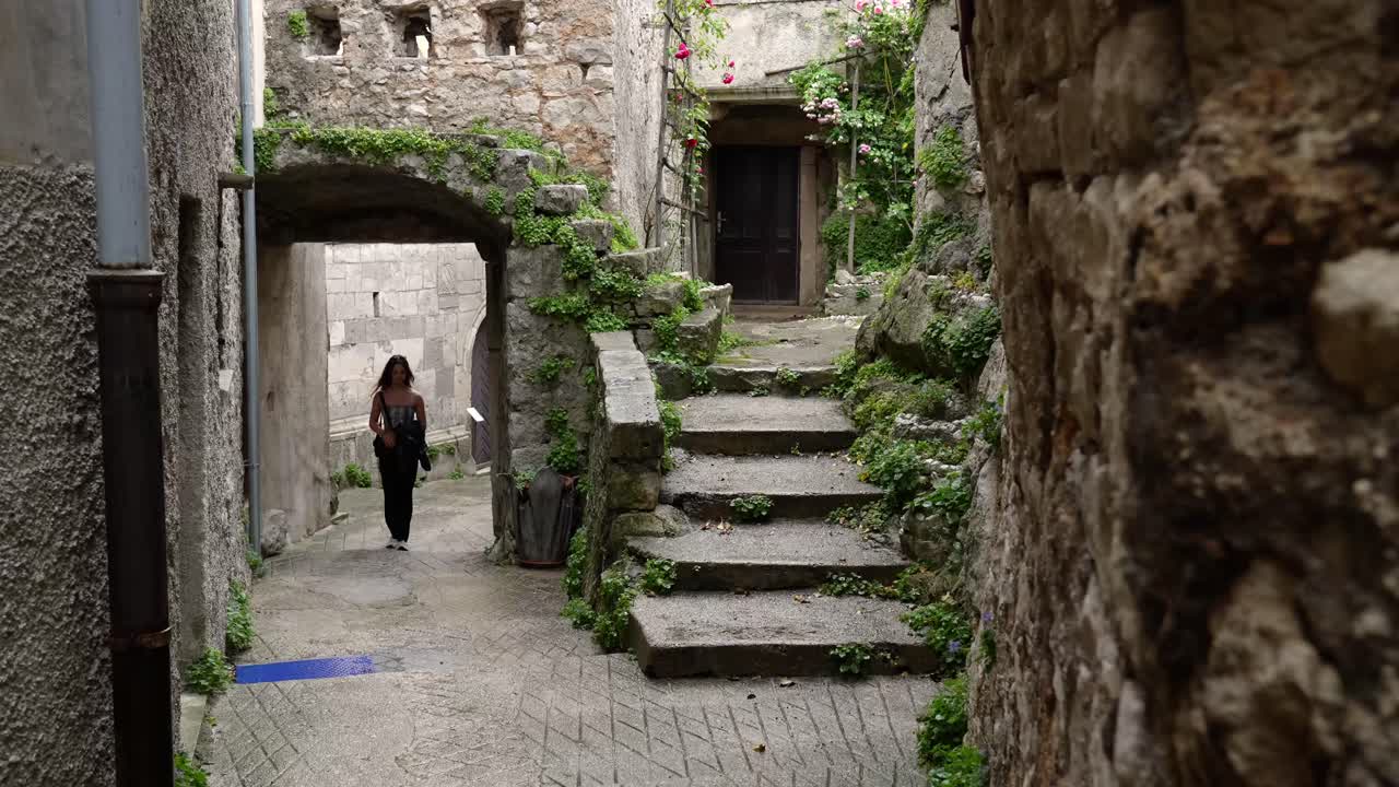 Giovane donna cammina attraverso un vicolo sotto un vecchio edificio in pietra ricoperto di vegetazione