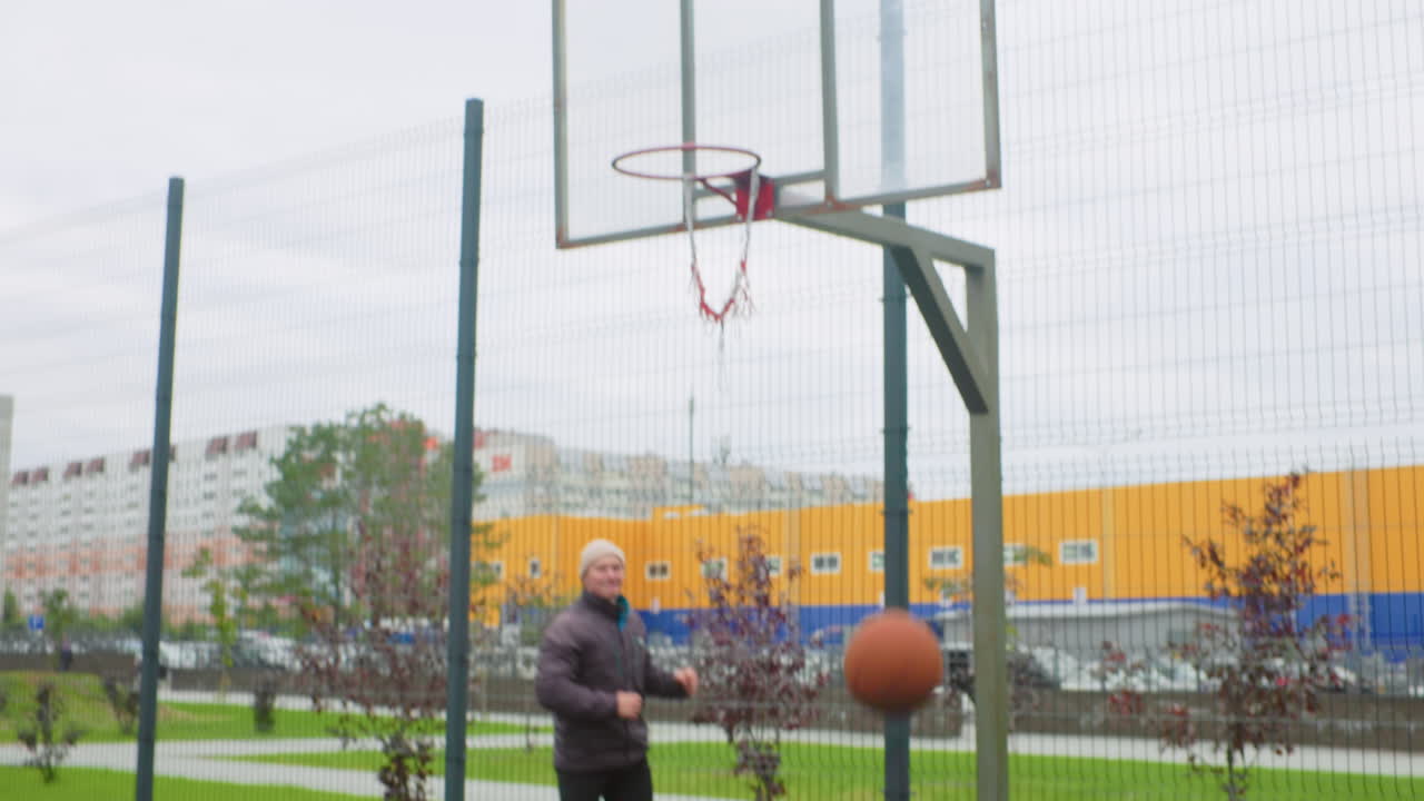 Uomo caucasico palleggia con un pallone da basket sul campo durante un allenamento solitario all'aperto, concentrato sul gioco di gambe, crossover controllato, tiri con rimbalzo verso il canestro, superficie in gomma ruvida, scarpe da ginnastica blu, abbigliamento casual