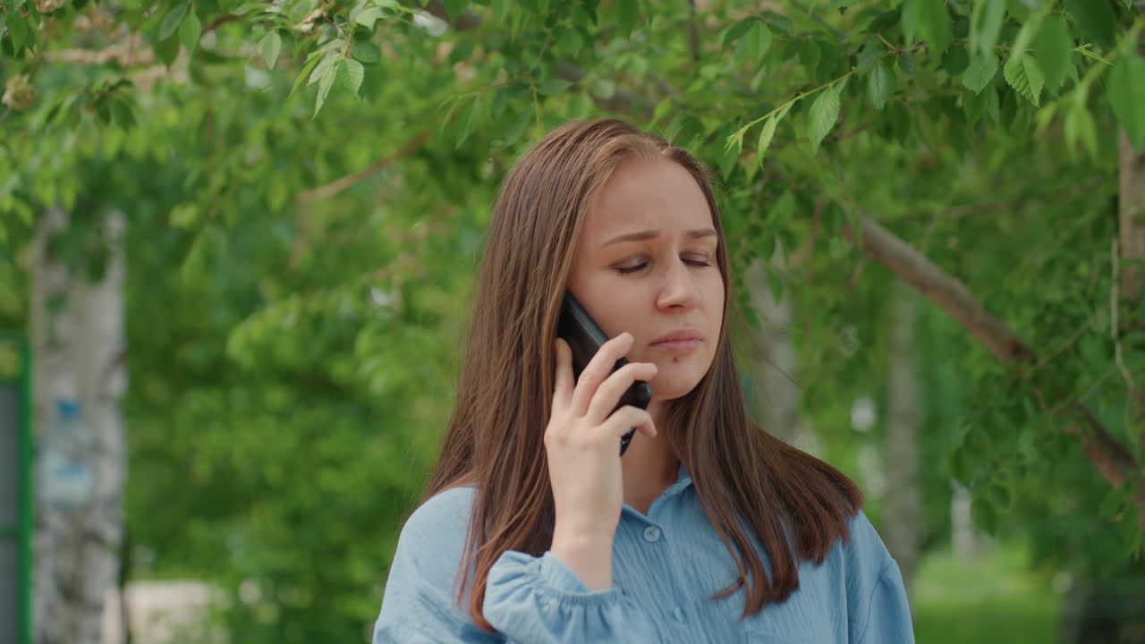 햇빛이 비치는 공원의 자작나무 옆에서 휴대전화를 들고 서 있는, 걱정스러운 표정의 백인 여성. 데님 셔츠를 입고 있으며, 손에 든 모바일로 대화 중. 미묘한 불안감, 기다리는 자세, 자연스러운 녹색 잎.