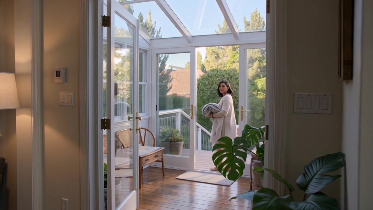 Donna che cammina, indossando un cardigan beige, si dirige verso la veranda per controllare il giardino, si ferma e raccoglie un asciugamano piegato