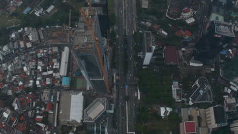 Toma-Aérea-De-Carro-Inclinándose-En-Una-Toma-Cenital-De-Un-Alto-Rascacielos-De-Vidrio-En-Construcción-Por-Una-Autopista-Concurrida