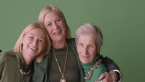 two blonde mature women and blonde senior woman smiling and hugging, wearing green tones clothes and posing on green background 1