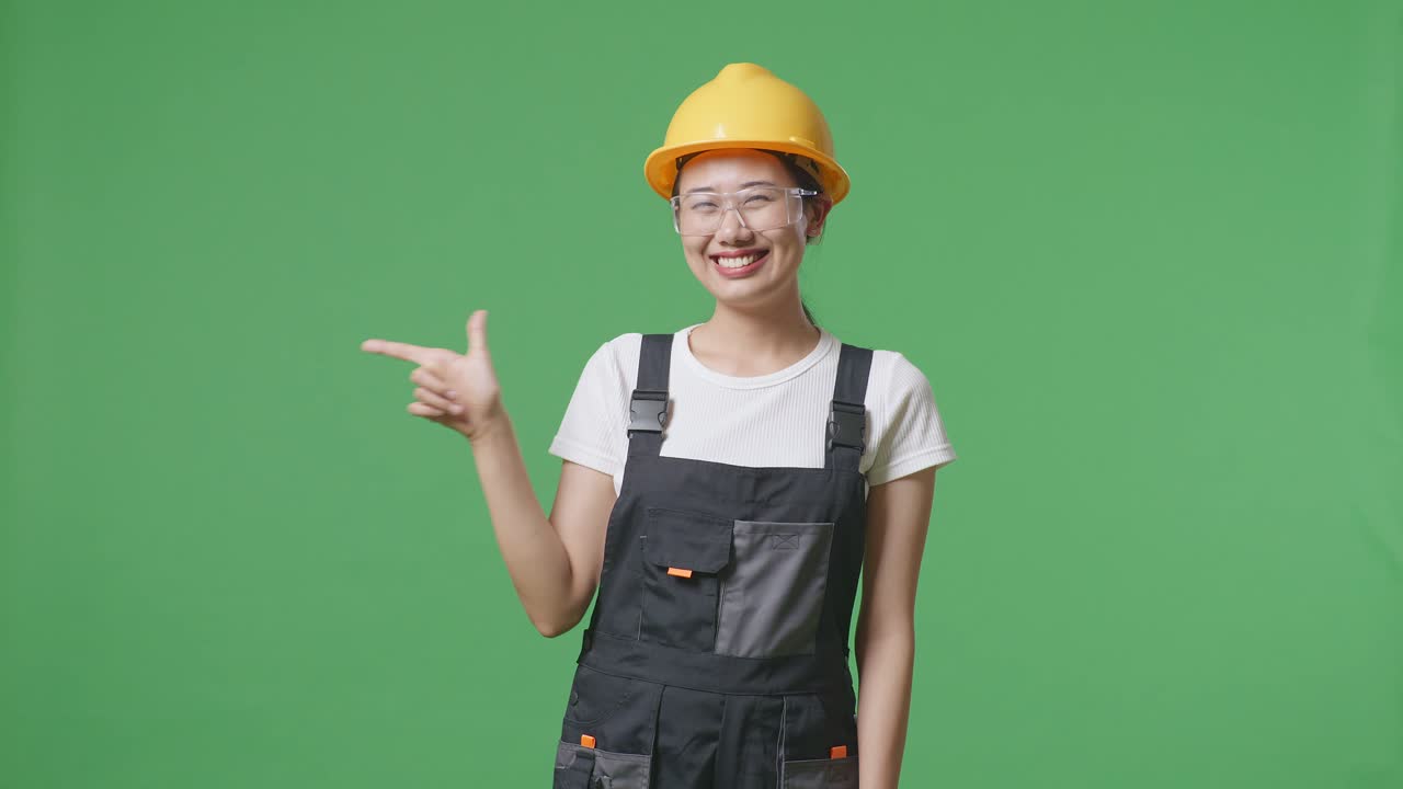 안경과 안전 헬을 입은 아시아 여성 노동자가 녹색 스크린 배경 스튜디오에 서서 웃고 을 가리키고 있습니다.