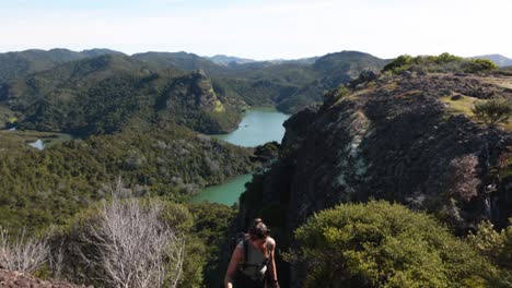 young caucasian woman climbing up a rock holding on to a ferrata rail with a view of a bay with mountains, greenery and turquoise water on a sunny day in duke's nose, northland, new zealand.