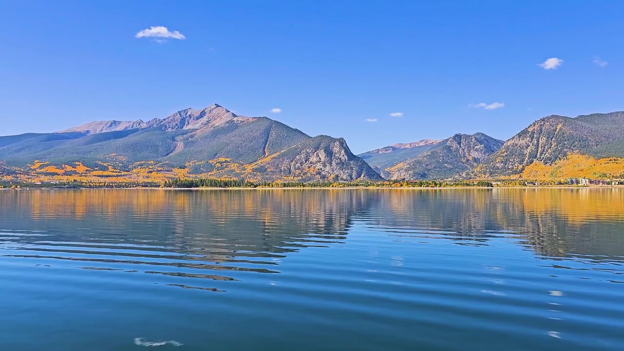 Ampia scena all'aperto a Dillon, Colorado, che mostra una vista panoramica di un brillante fogliame autunnale, pendii montuosi boscosi, superficie calma del lago, riva sabbiosa, cielo azzurro limpido e paesaggio alpino.